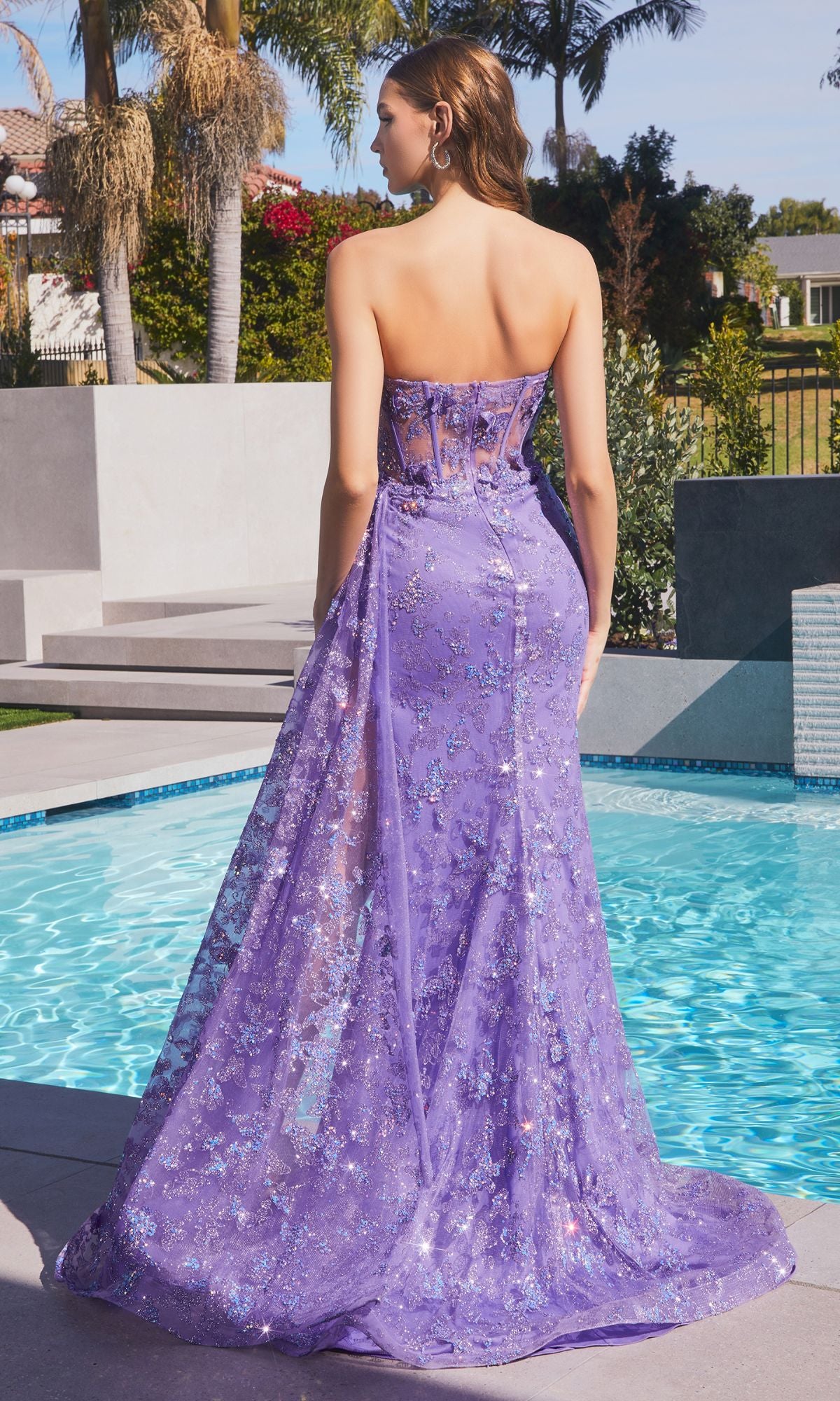 A woman stands by a pool in the Strapless Long Butterfly Prom Dress CD998, a floor-length purple sequin gown with a sheer overlay, facing away from the camera.